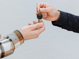 Two hands exchanging a key, with one holding documents. One person wears a plaid shirt; the other, a black sleeve. Neutral background.