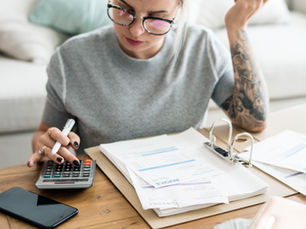 Woman with glasses and tattoos calculating invoices using a calculator at a wooden table. Papers, binder, and smartphone nearby.