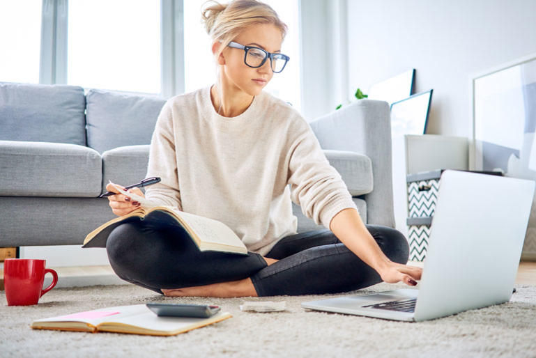 Woman in glasses sitting on carpet with laptop, holding a book and pen. Grey couch, red mug, notepad, and calculator nearby. Bright room.