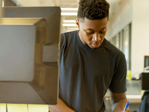 Man writing in a notebook at a desk in an office, focused and concentrated. Computer, keyboard, and sticky notes visible. Bright lighting.
