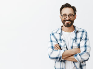 Smiling man with glasses and beard in a blue plaid shirt, arms crossed, standing against a white background. Relaxed and happy mood.