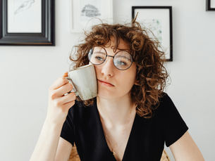 Curly-haired person with glasses, holding a mug to their cheek, looking contemplative. Frames on white wall in background.