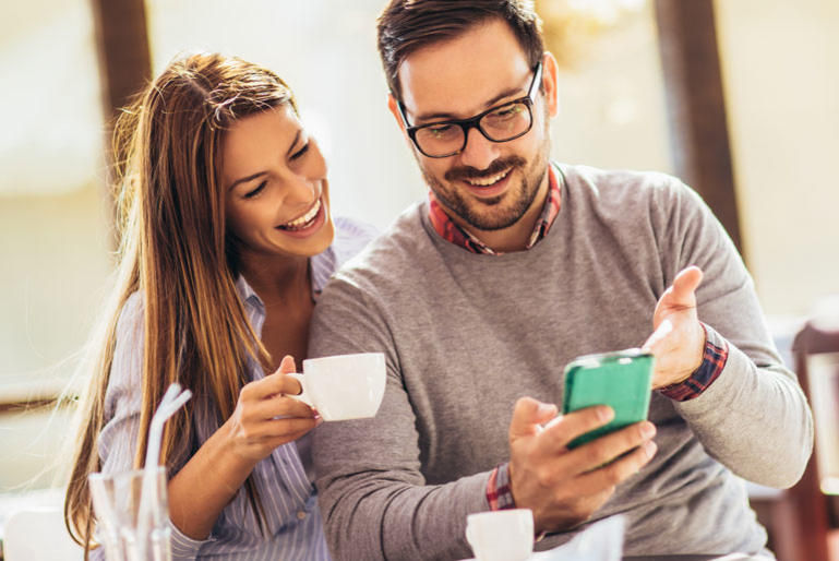 A smiling man and woman sit in a bright room, looking at a phone. The woman holds a coffee cup. Both appear happy and engaged.