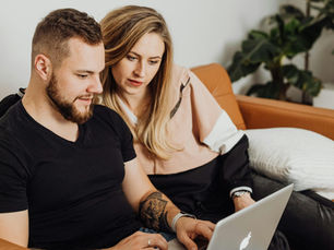 Couple on a brown sofa using a laptop. The man in a black shirt is typing, while the woman in a pink and black top looks on. A plant is in the background.