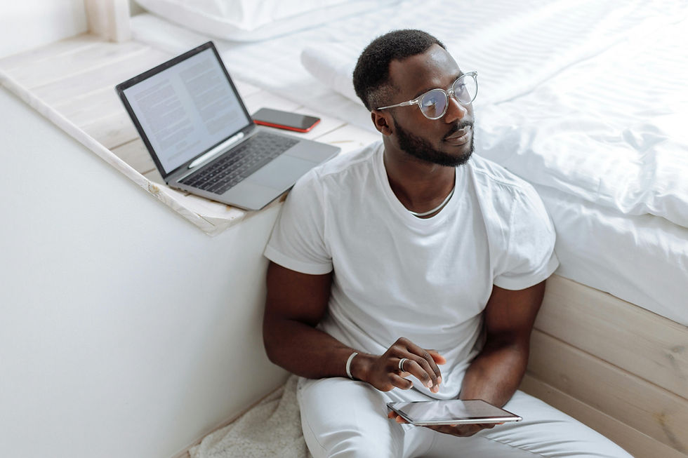 Man in white sitting against a bed, holding a tablet. Laptop and phone beside him. Calm expression, white bedding background.