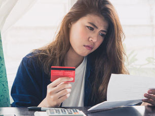 A woman looks concerned, holding a red credit card and a paper, sitting at a table with a calculator.