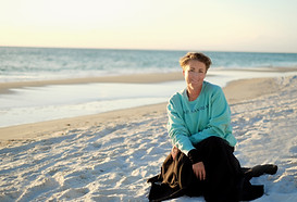Elizabeth Hockmeyer in a giant black ballroom skirt, teal oversized sweatshirt, crouched on a white sand beach at sunset.