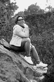Woman sitting on rock, smiling, wearing glasses and casual clothing outdoors.