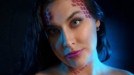 Woman with scale makeup, gazing forward with artistic blue and purple lighting.