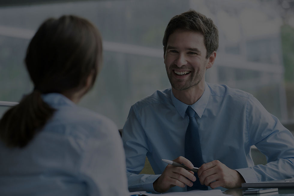 Two people in business attire talking at a desk. The man smiles, holding a pen. Bright office with large windows in the background.