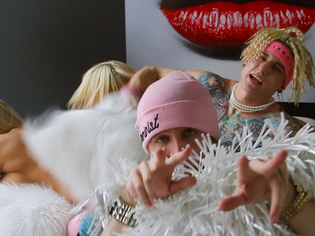 People with blonde hair pose energetically in a room. One wears a pink beanie, another has jewelry. A large red lips painting is behind them.