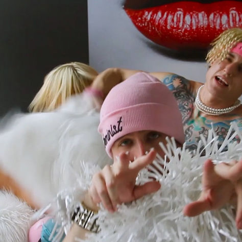 People with blonde hair pose energetically in a room. One wears a pink beanie, another has jewelry. A large red lips painting is behind them.