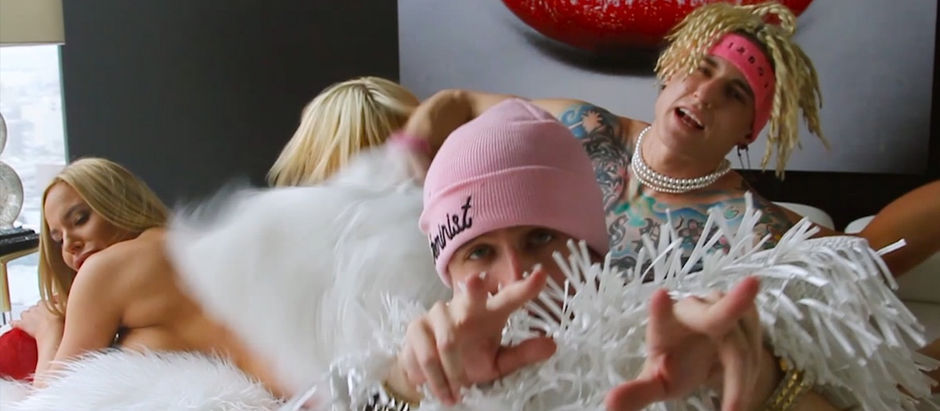 People with blonde hair pose energetically in a room. One wears a pink beanie, another has jewelry. A large red lips painting is behind them.