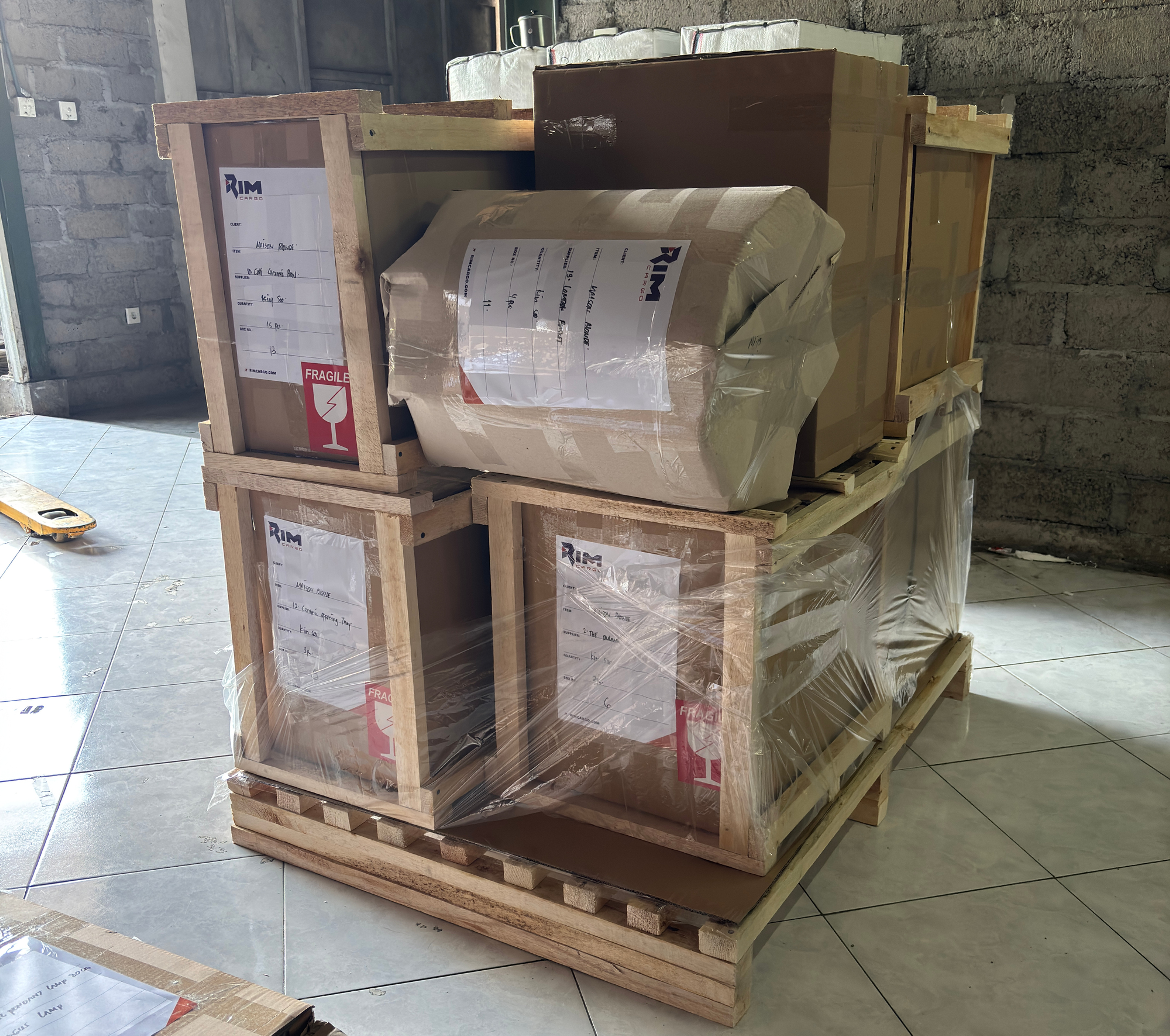 Stacked wooden shipping crates and cardboard boxes wrapped on a pallet in an unfinished room, with fragile labels and dim light