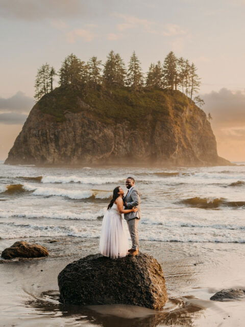 A bride and groom stand on a rock by the shore.
