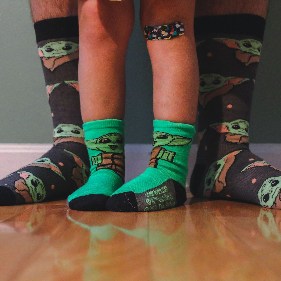 Two pairs of legs: child with green "Baby Yoda" socks and band-aid, adult with matching socks. Standing on wooden floor, green wall backdrop.