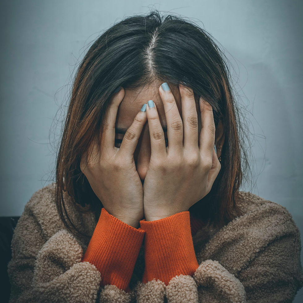 A person covers their face with their hands, showing light blue nail polish. They wear a brown coat with orange cuffs; background is gray.