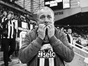 A powerful, medium close-up shot of a distraught female Newcastle United supporter at the end of the match. She is standing near the front of the Gallowgate End, her iconic black-and-white striped "Howe's Geordie Army" scarf twisted around her neck. Her hands are pressed firmly against her face, palms digging into her cheeks, and her mouth is open in a silent gasp of anguish. Her eyes, filled with genuine terror, are locked on the St. James' Park big screen, which prominently displays a generic Premier League 'Bottom of the Table' graphic showing Newcastle in 18th (or a stylized 'Relegation Zone' visual). The late afternoon sun highlights the sweat on her forehead and the blurred, desolate faces of other fans leaving the stadium around her