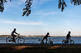 Family cycling and scootering along a lake under blue sky.
