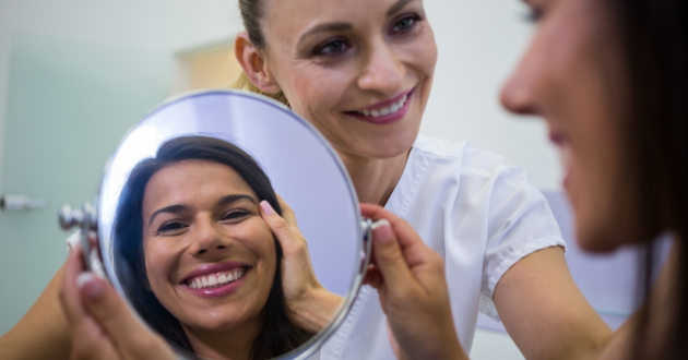 Woman smiles while holding a round mirror showing her reflection. Another woman in a white coat smiles beside her. Bright, cheerful setting.
