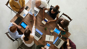 A diverse group of eight people sit around a wooden table in a collaborative meeting. Two individuals in the center shake hands, symbolizing partnership. Laptops, tablets, notebooks, coffee cups, and small plants are on the table, with screens displaying the word ‘PROJECT,’ highlighting teamwork and professional collaboration.