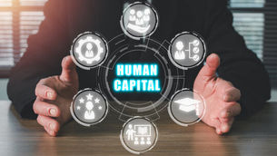 Business person at a desk interacting with a digital interface displaying “HUMAN CAPITAL” surrounded by icons of people, education, performance ratings, and organizational structures, symbolizing workforce value, skills, and productivity.