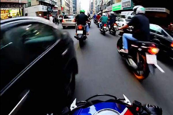 Motociclistas trafegando em via urbana com fluxo intenso e comportamento de risco — imagem capturada de dentro de outra moto mostra proximidade perigosa com carro, ilustrando causas comuns de acidentes como velocidade, imprudência e ultrapassagens indevidas.