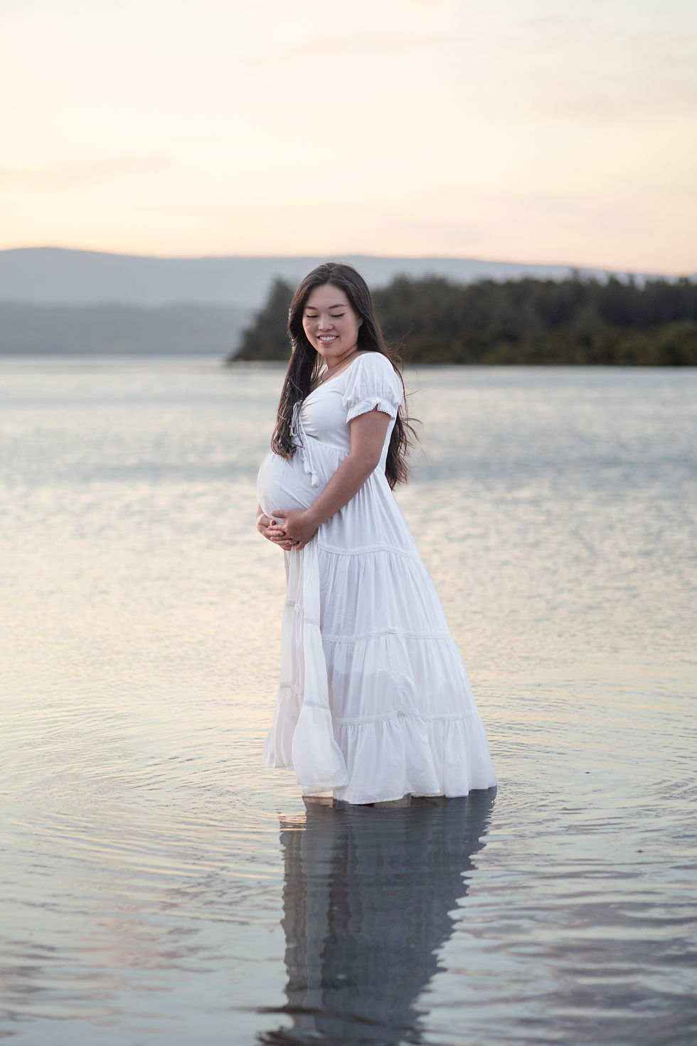First time mum standing in a white dress in the middle of a calm lake during maternity photos
