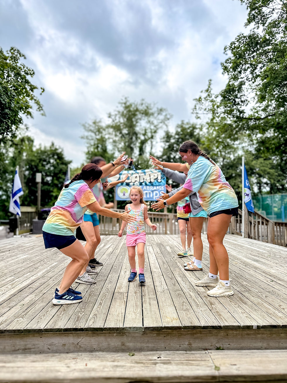 Adults in tie-dye shirts cheer as a child runs between them on a wooden deck. Camp sign in background. Trees and flags visible. Festive mood.