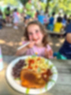Child joyfully eating pasta outdoors at a picnic, with blurred kids in the background. Plate includes beans and toast. Warm, sunny day.