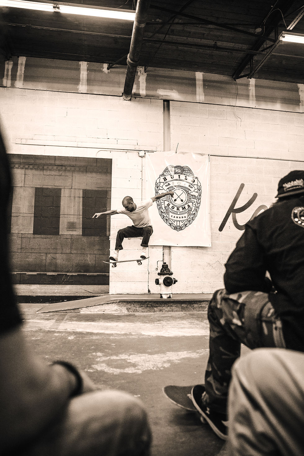 Skateboarder midair in an indoor skate park with seated onlookers in foreground.