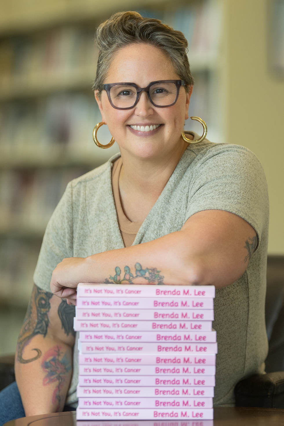 A woman with dimples, glasses, and salt and pepper hair leaning on a stack of books titled "It's not you, It's Cancer".
