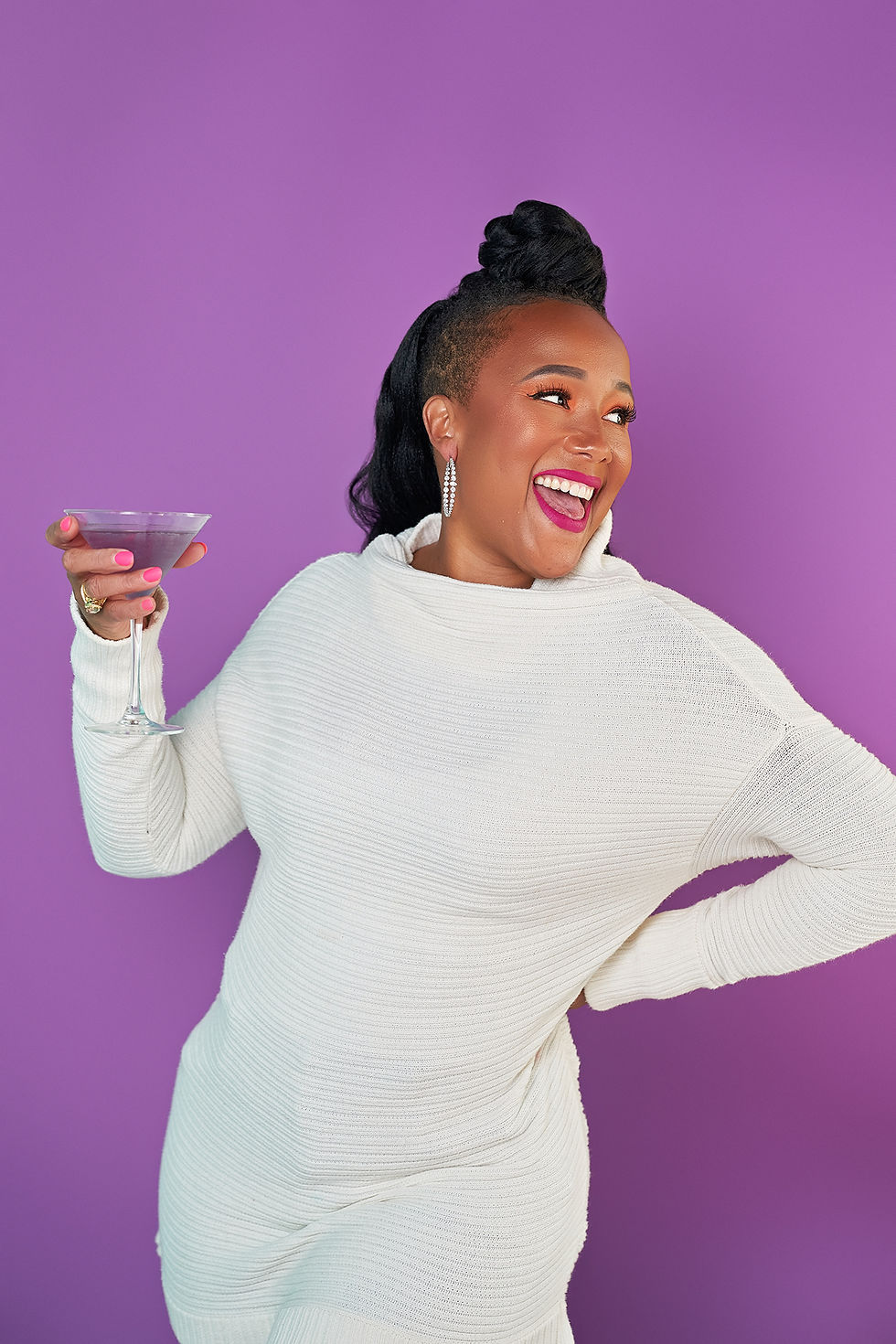 Woman in a white sweater holding a cocktail glass, laughing against a purple background. She has bright pink lipstick and earrings.