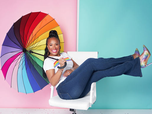 Smiling woman in jeans and rainbow heels, holding a rainbow umbrella, sits on a white chair against a pink and teal background.