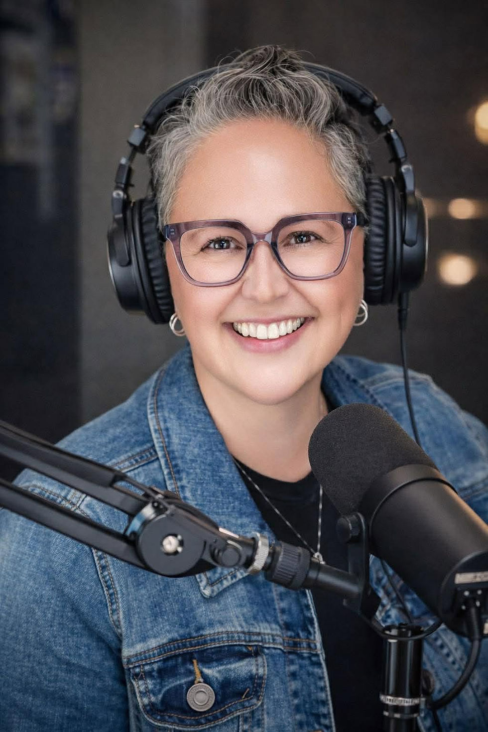 A woman with glasses, salt-n-pepper hair, and a denim jacket with headphone on and a microphone.