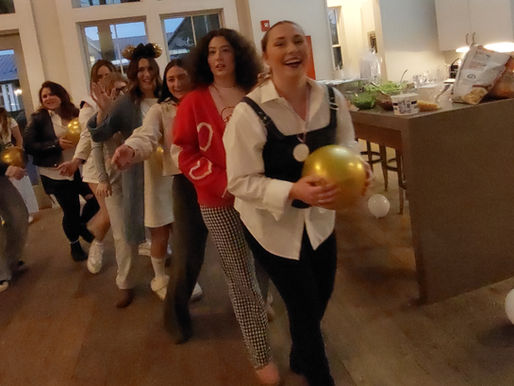 People in a conga line holding gold balloons, smiling indoors with a warm, festive atmosphere. Snacks and drinks on the table nearby.