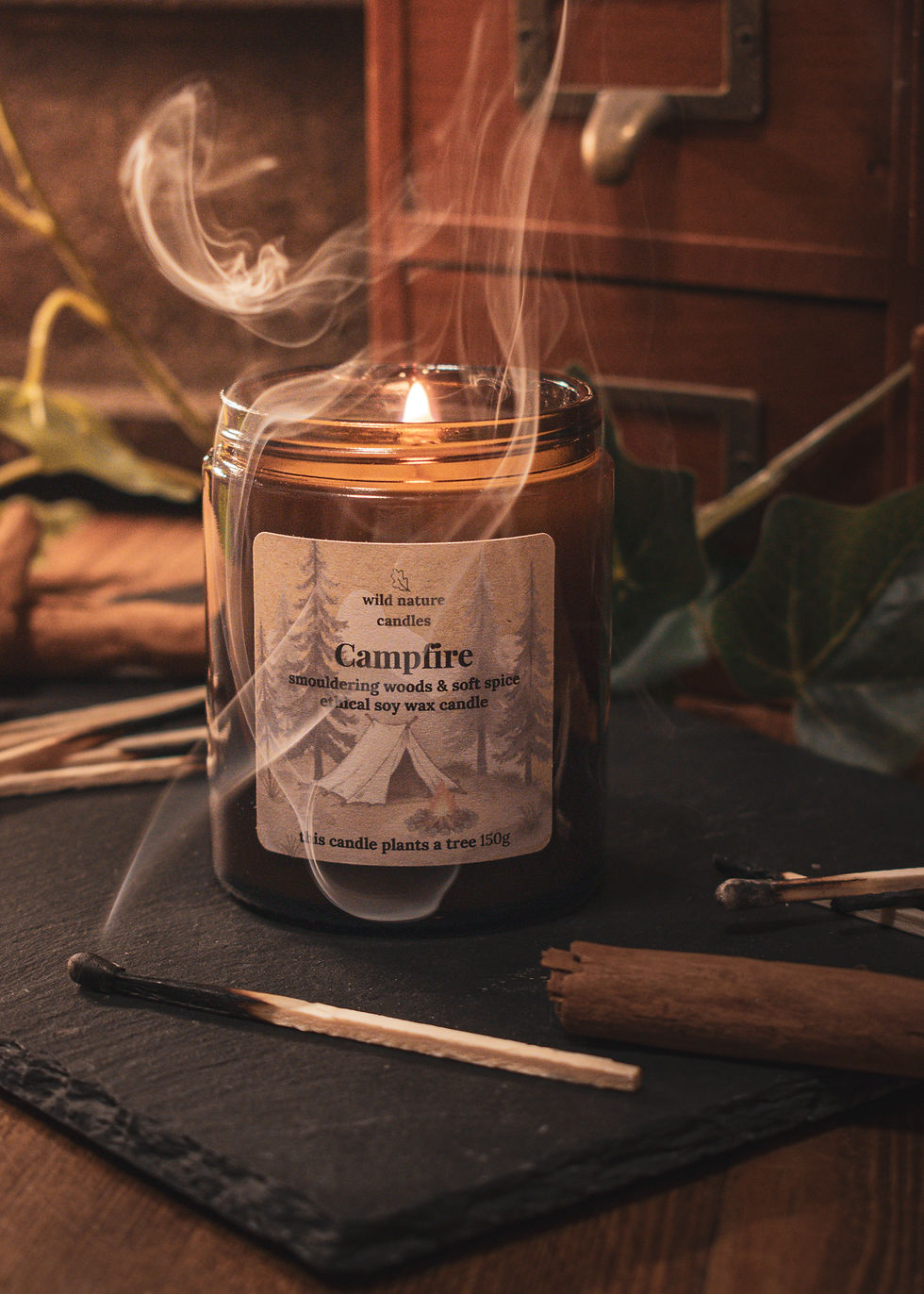 Campfire soy candle burning with smokey match & curling smoke, styled on black slate with burnt matches and cinnamon for a c