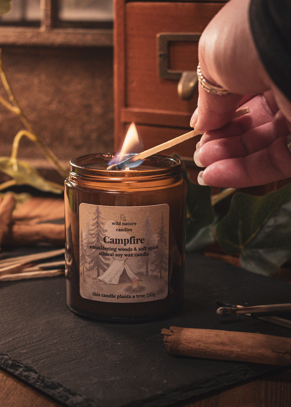 Campfire soy candle being lit on black slate with burnt matches and cinnamon sticks, warm woodland-inspired home fragrance