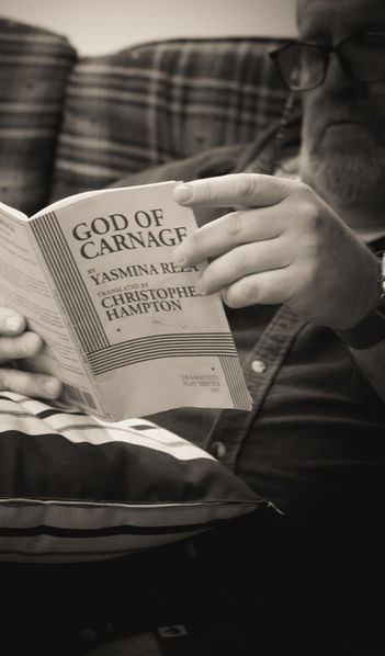 A photo from rehearsals for the play, "God of Carnage." One of the actors sits and reads the script, resting it on a pillow.