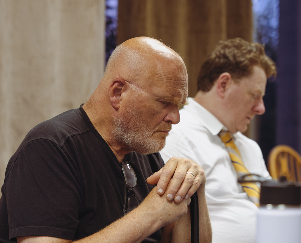 A photo from rehearsals for the play "12 Angry Men". An image of two men sitting at a table. The man in the foreground has his hands crossed and his head is bowed.