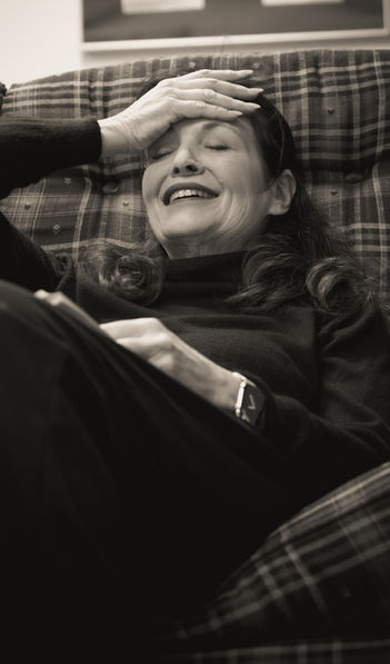 A photo from rehearsals for the play, "God of Carnage." A woman sits on a couch, laughing and pressing the palm of her hand to her forehead.