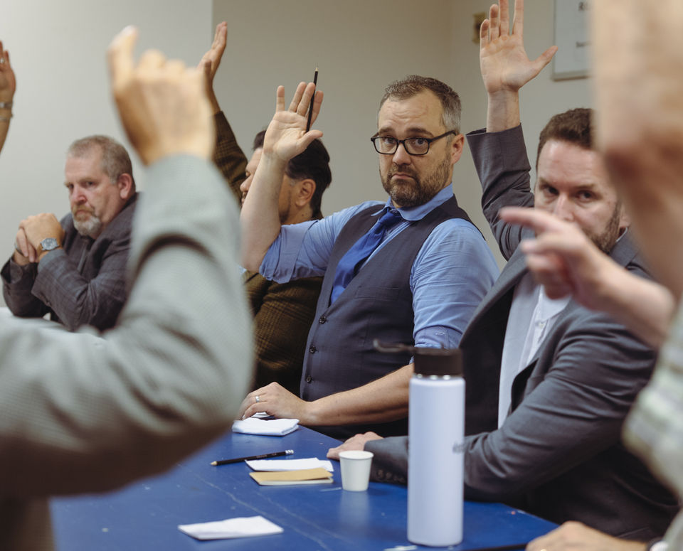 A photo from rehearsals for the play "12 Angry Men". Men in suits sit around a table, raising their hand. One man abstains from raising his hand.