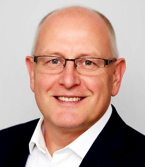 John Swain wearing a white shirt and blue suit, smiling