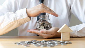 Person in white shirt, black tie holds a jar of coins on a table with scattered coins and a small wooden house. Emphasizes saving money.