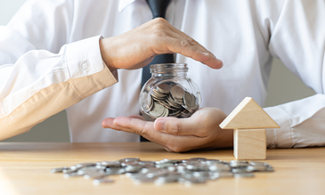 Person in white shirt, black tie holds a jar of coins on a table with scattered coins and a small wooden house. Emphasizes saving money.