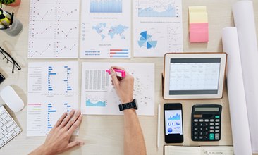Hands highlighting charts with pink marker on desk. Graphs, tablet with calendar, smartphone, calculator, and stationery scattered around.