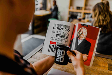 Person in a coffee shop reads a magazine titled "The Entrepreneurs Guide to Success & Business Growth." Holding a black "80/20" mug.