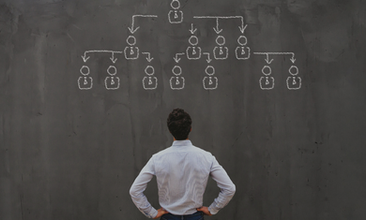 Person in striped shirt looks at a chalk-drawn organizational chart on a dark chalkboard. Atmosphere is thoughtful and focused.