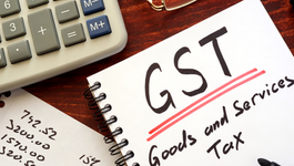 Calculator on desk next to notebook with "GST: Goods and Services Tax" written on it, along with handwritten numbers. Brown wood background.