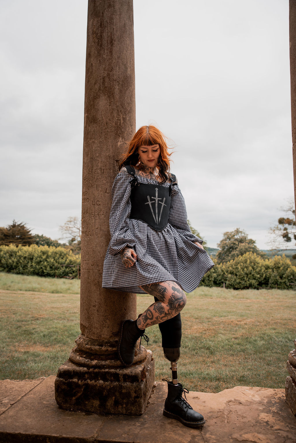 a tattooed model wearing a gingham dress with black corset leaning against a column